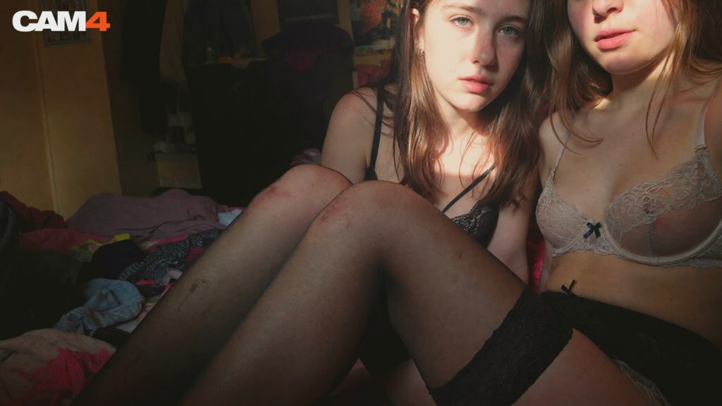 Sex Live en Couple : Deux jeunes femmes aux longs cheveux, assises côte à côte sur un lit en désordre, dans une chambre faiblement éclairée. L’une porte une lingerie noire avec bas résille, l’autre une lingerie en dentelle beige clair transparente. Ambiance intime et sensuelle, logo CAM4 en haut à gauche.