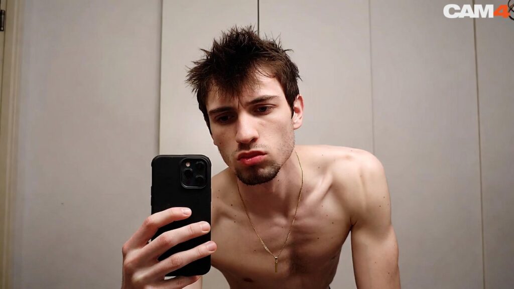 Un jeune homme torse nu aux cheveux bruns ébouriffés et barbe naissante, tenant un smartphone noir dans la main pour prendre un selfie miroir, portant une chaîne en or avec pendentif, regardant vers la caméra la bouche en moue. Logo CAM4 en haut à droite.