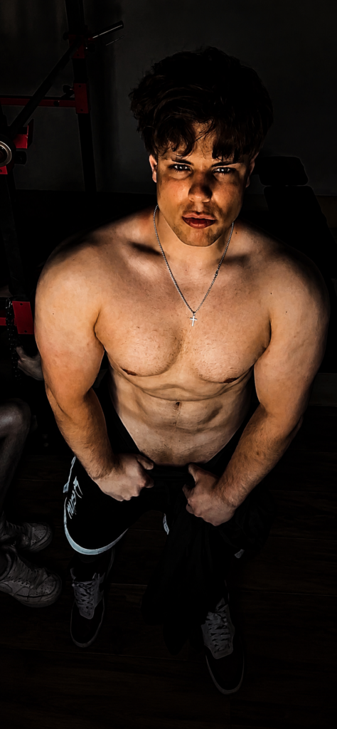 Homme musclé torse nu portant une chaîne avec croix, regardant l’appareil photo de bas en haut, mains sur son pantalon dans une salle de sport.